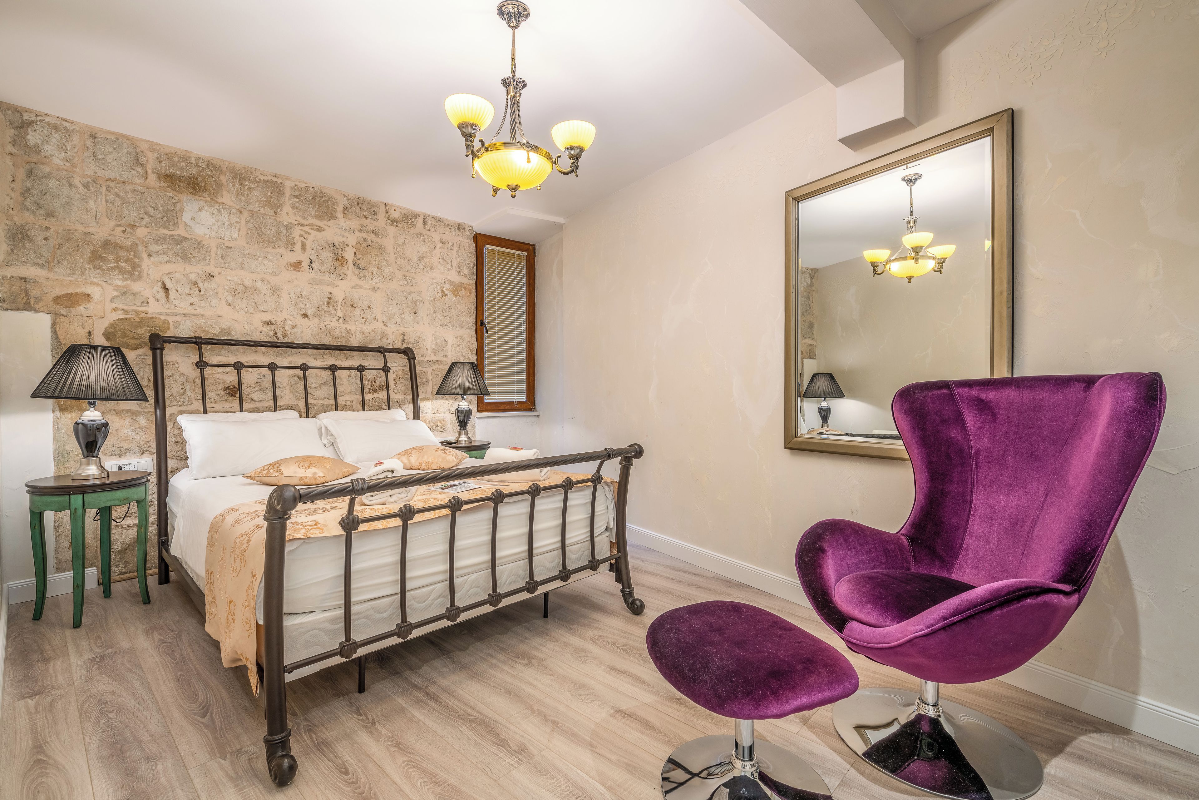 A bedroom with a stone wall, a bed with a headboard, a purple chair, a mirror, and a chandelier.