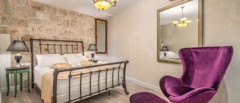 A bedroom with a stone wall, a bed with a headboard, a purple chair, a mirror, and a chandelier.