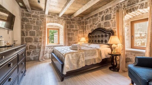 Ein Schlafzimmer mit Steinwänden, einem Bett mit Kopfteil, einem Schminktisch und einem Fenster mit Vorhängen.