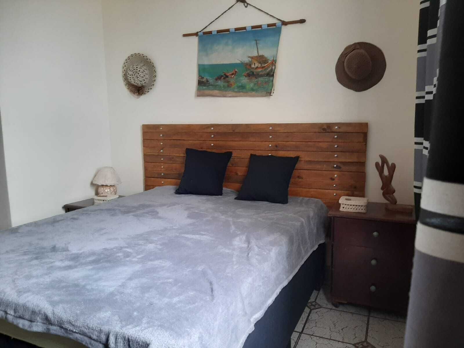A bedroom with a wooden headboard, two black pillows, a grey bedspread, a small bedside table with a lamp, a wall hanging, and a decorative item on the nightstand.