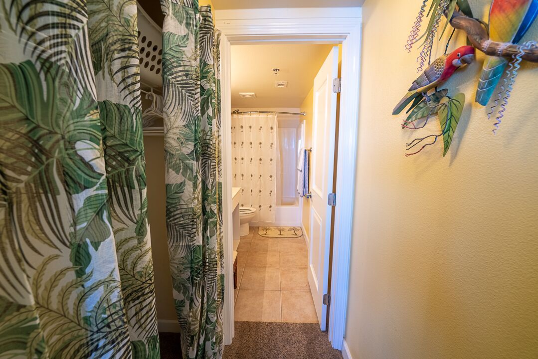 A narrow hallway with a shower curtain featuring a tropical leaf pattern, leading to a bathroom with a toilet and a shower curtain with a floral pattern.