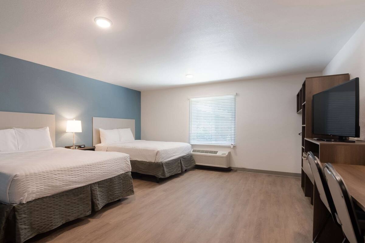 A hotel room with two beds, a TV, a desk, and a window with blinds.