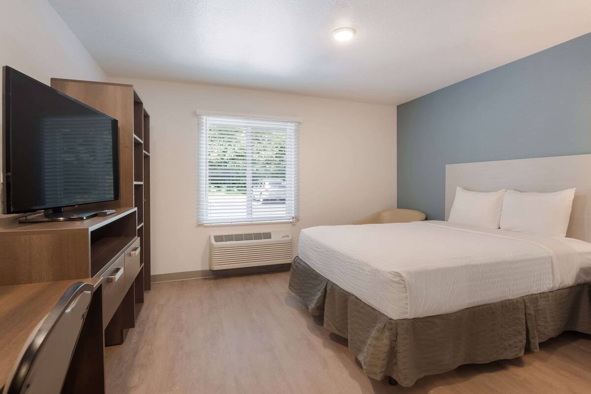 A hotel room with a bed, a TV, a desk, and a window with blinds.