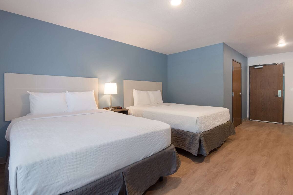 A hotel room with two beds, a nightstand with a lamp, a wooden floor, and a door.