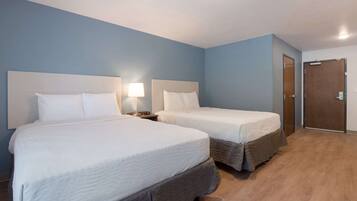 A hotel room with two beds, a nightstand with a lamp, a wooden floor, and a door.