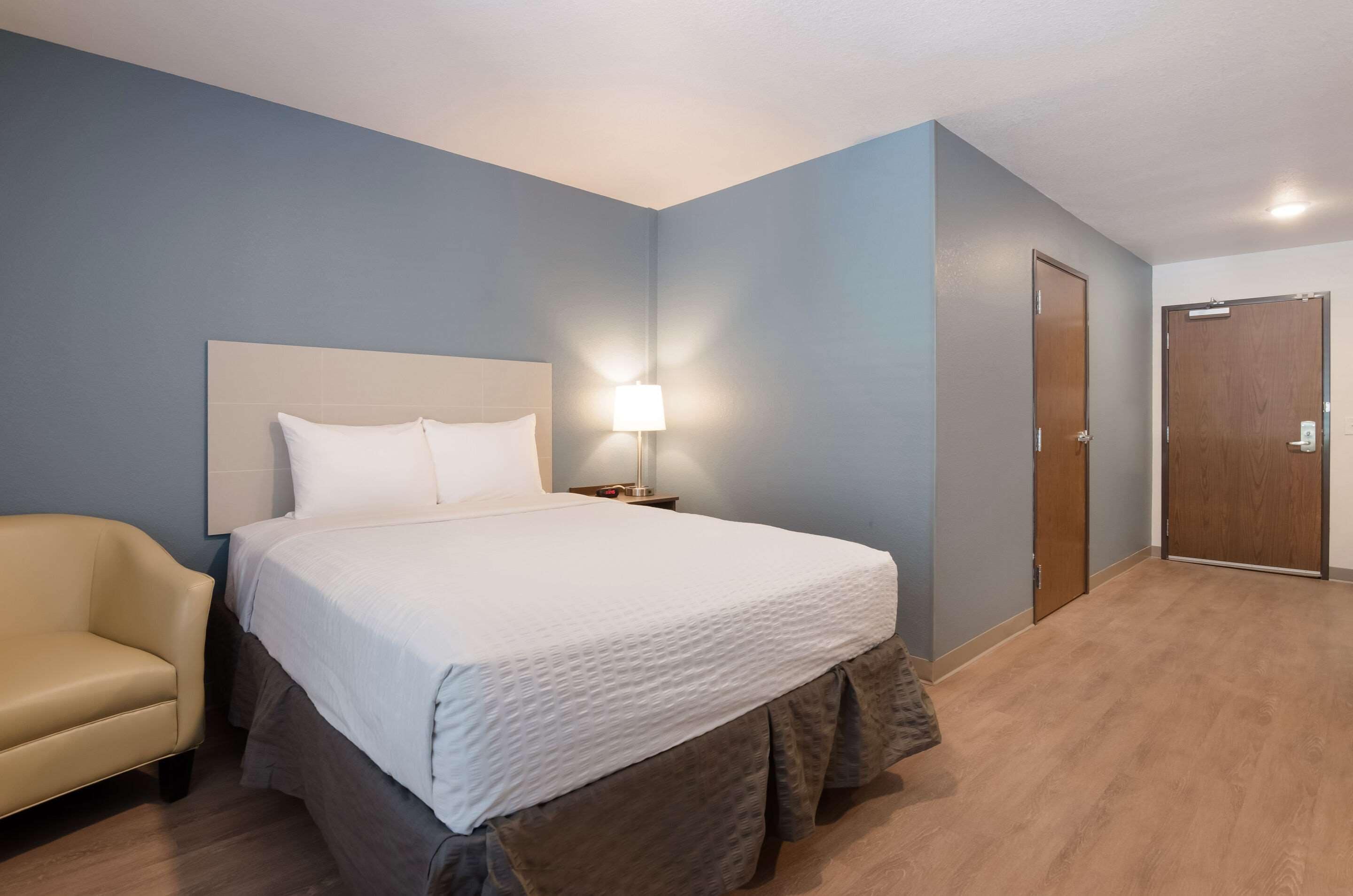 A hotel room with a bed, a nightstand, a lamp, a chair, and two doors.