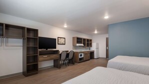 Ein Hotelzimmer mit einer Küchenzeile, einem Schreibtisch mit Stühlen, einem Fernseher und zwei Betten.