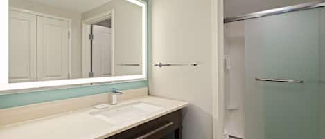 Un baño con un espejo grande, un lavamanos, un perchero para toallas y una cabina de ducha con mampara de vidrio.