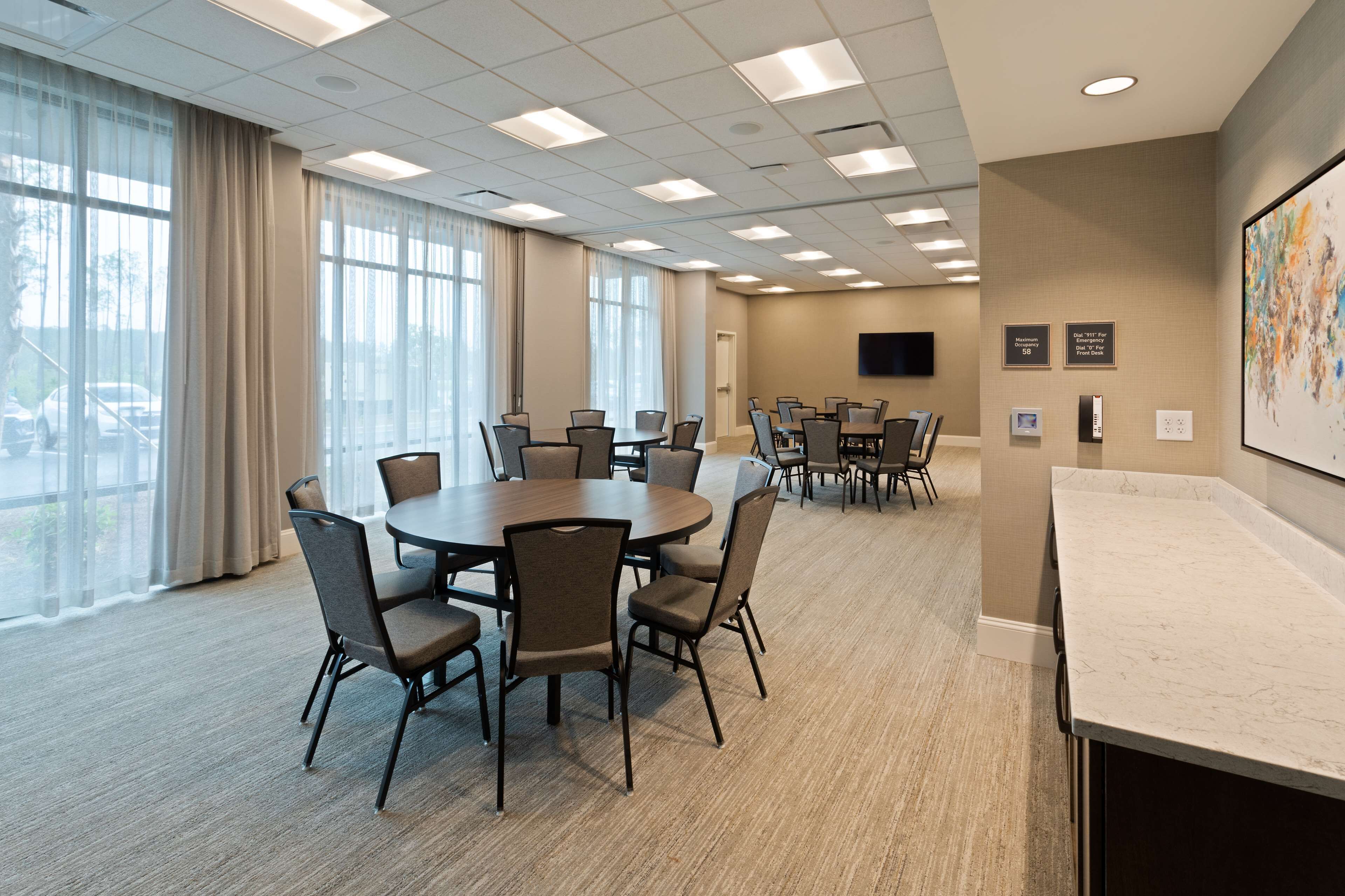 A conference room with a large table and chairs, a flat-screen TV on the wall, and a view of parked cars through large windows.