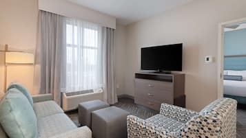 A hotel room with a sofa, ottoman, TV, and a bed with a blue headboard.
