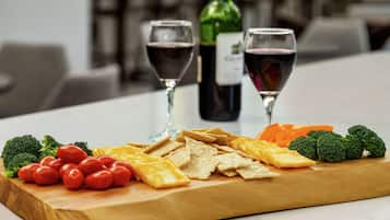 Una tabla de madera con queso, galletas y verduras. Se ven dos copas de vino tinto al fondo.