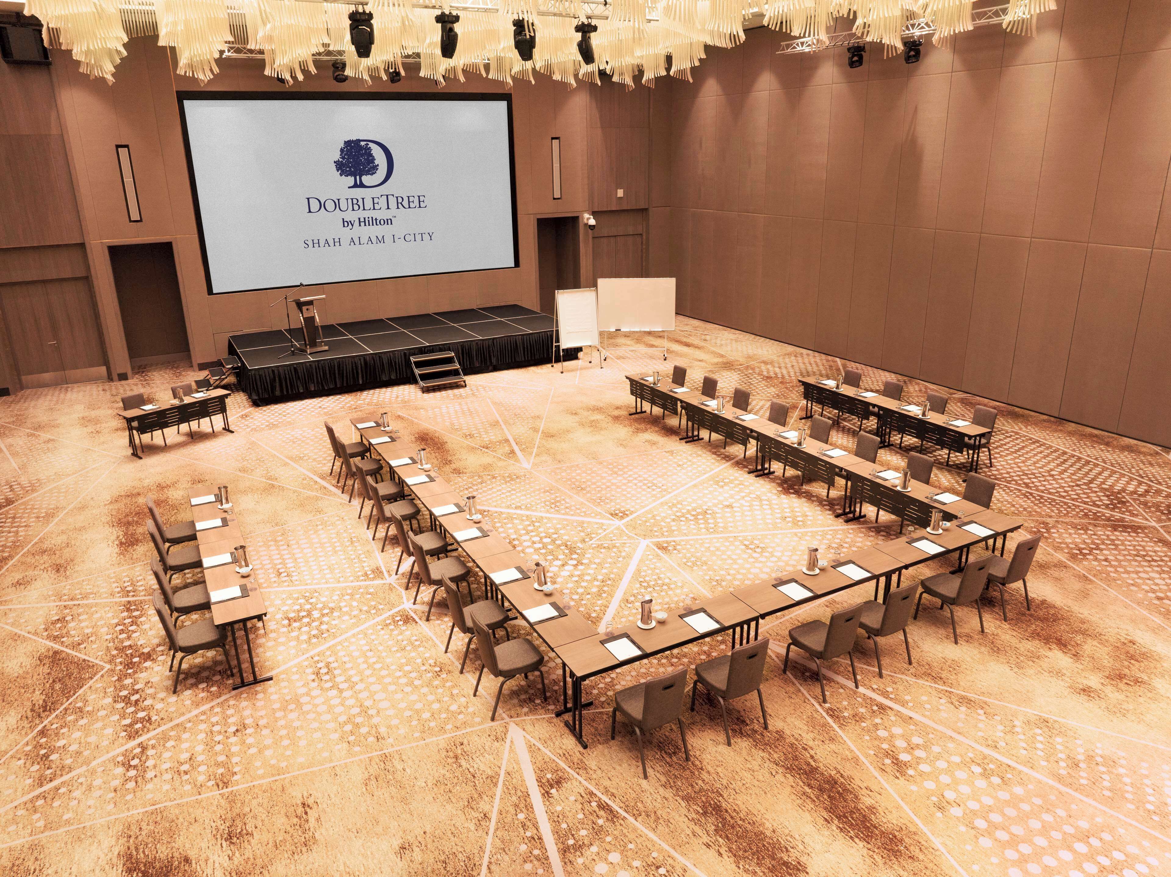 A conference room with a stage, multiple tables, and chairs, and a large screen displaying the DoubleTree by Hilton logo.