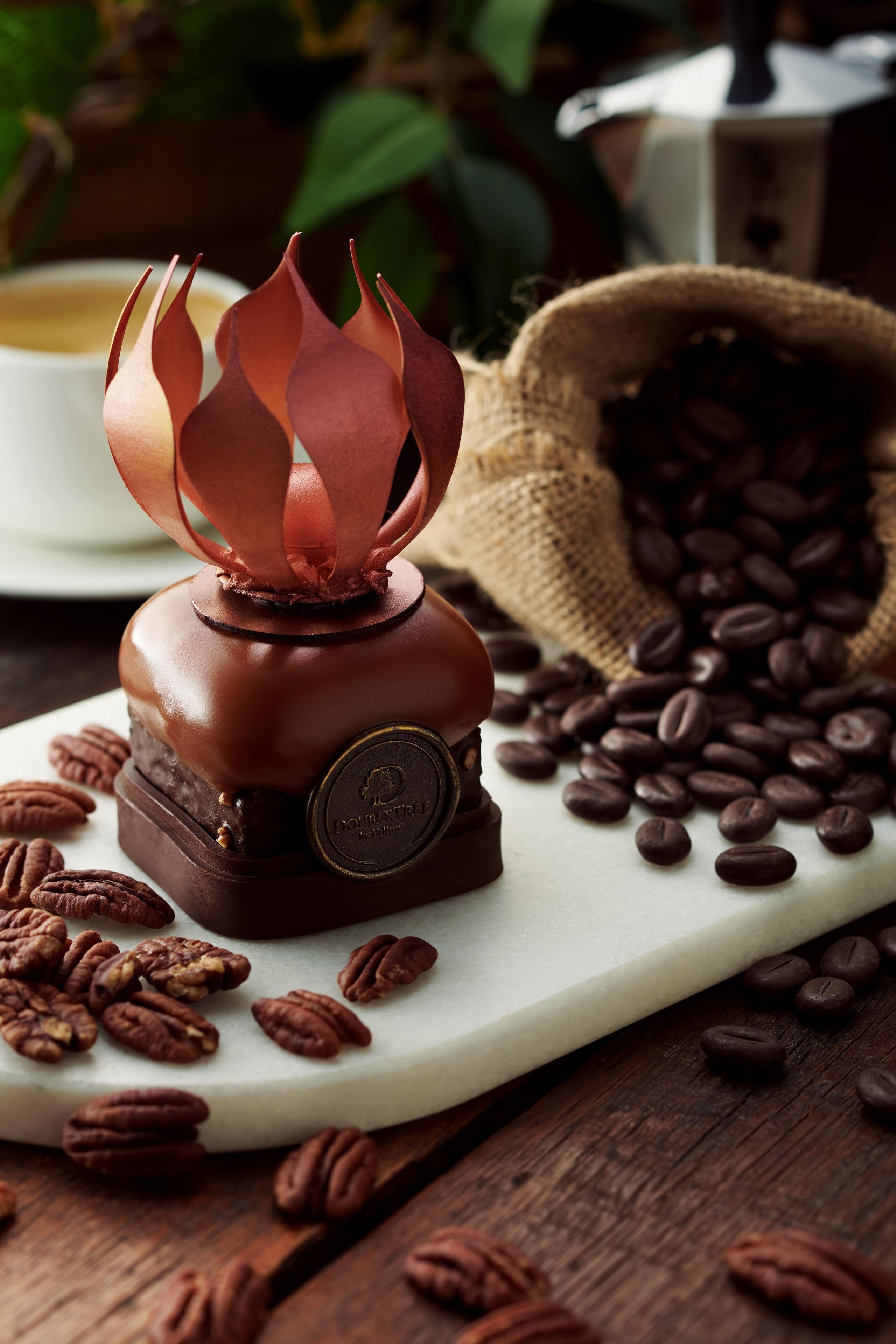 A coffee roaster with a flame design on top, surrounded by coffee beans and pecans on a white plate.
