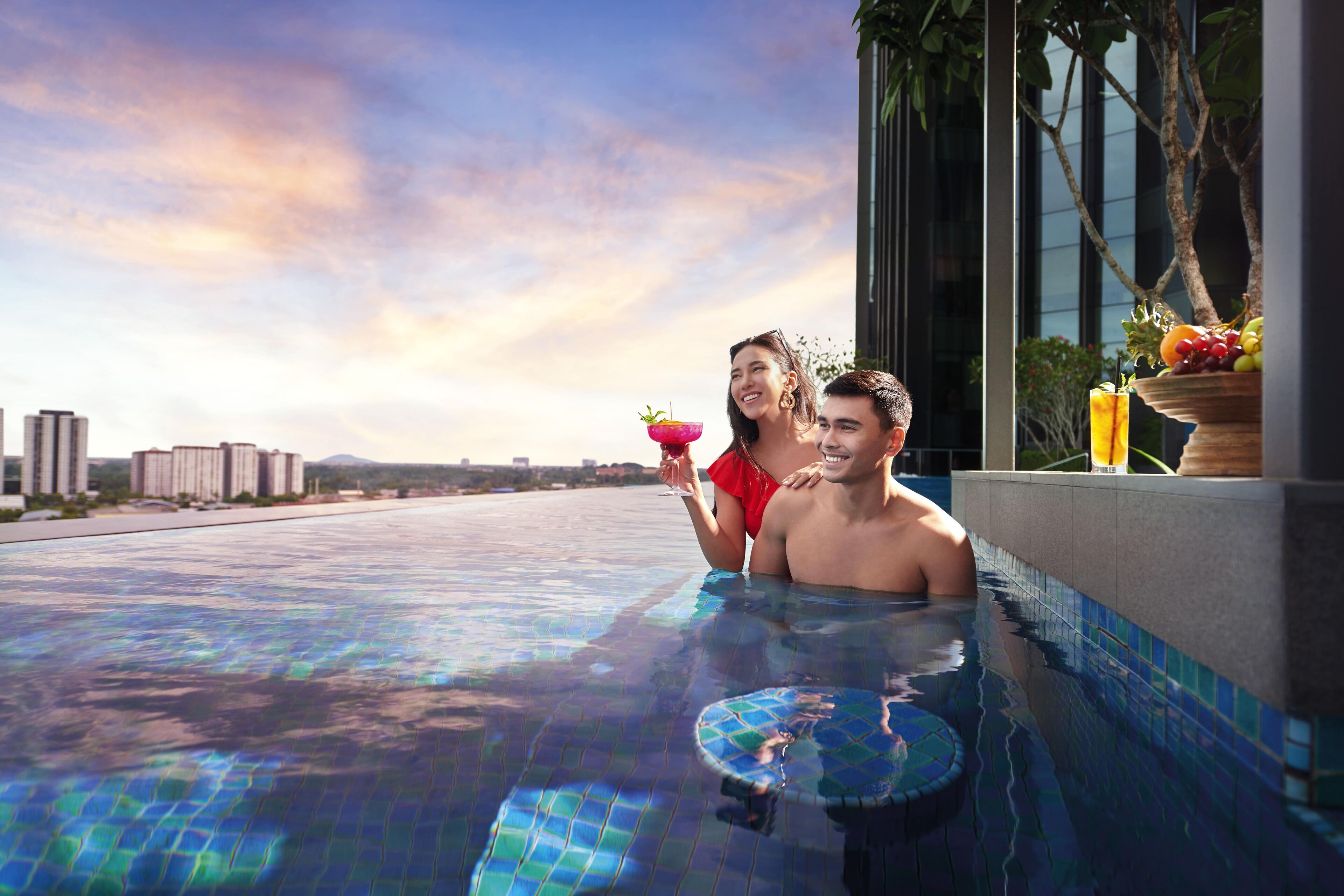 A couple in a rooftop infinity pool with a city view, a fruit bowl, and a drink on the side.