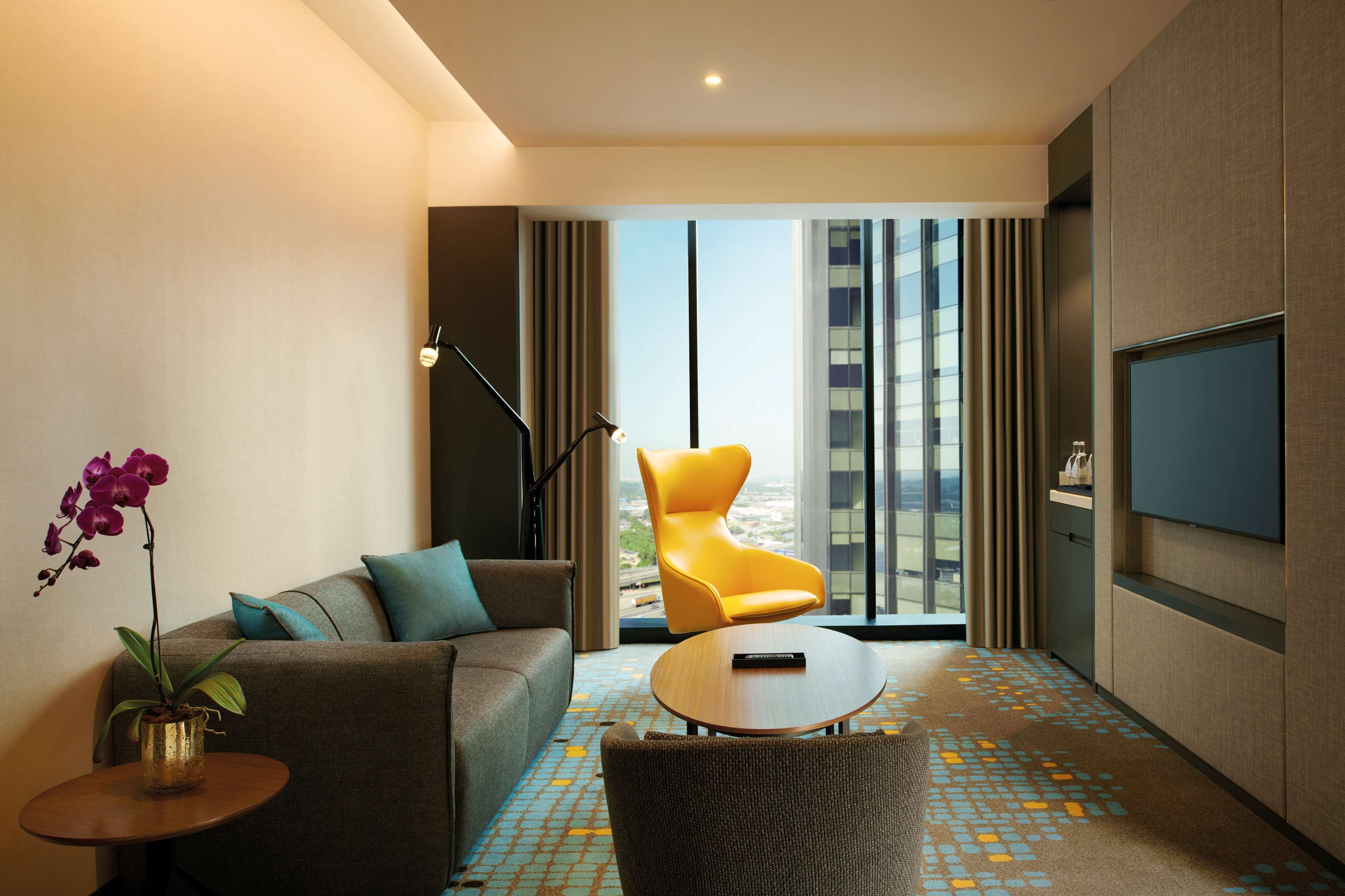 A modern hotel room with a sofa, a yellow armchair, a small round table, a TV, and a view of the city through large windows.