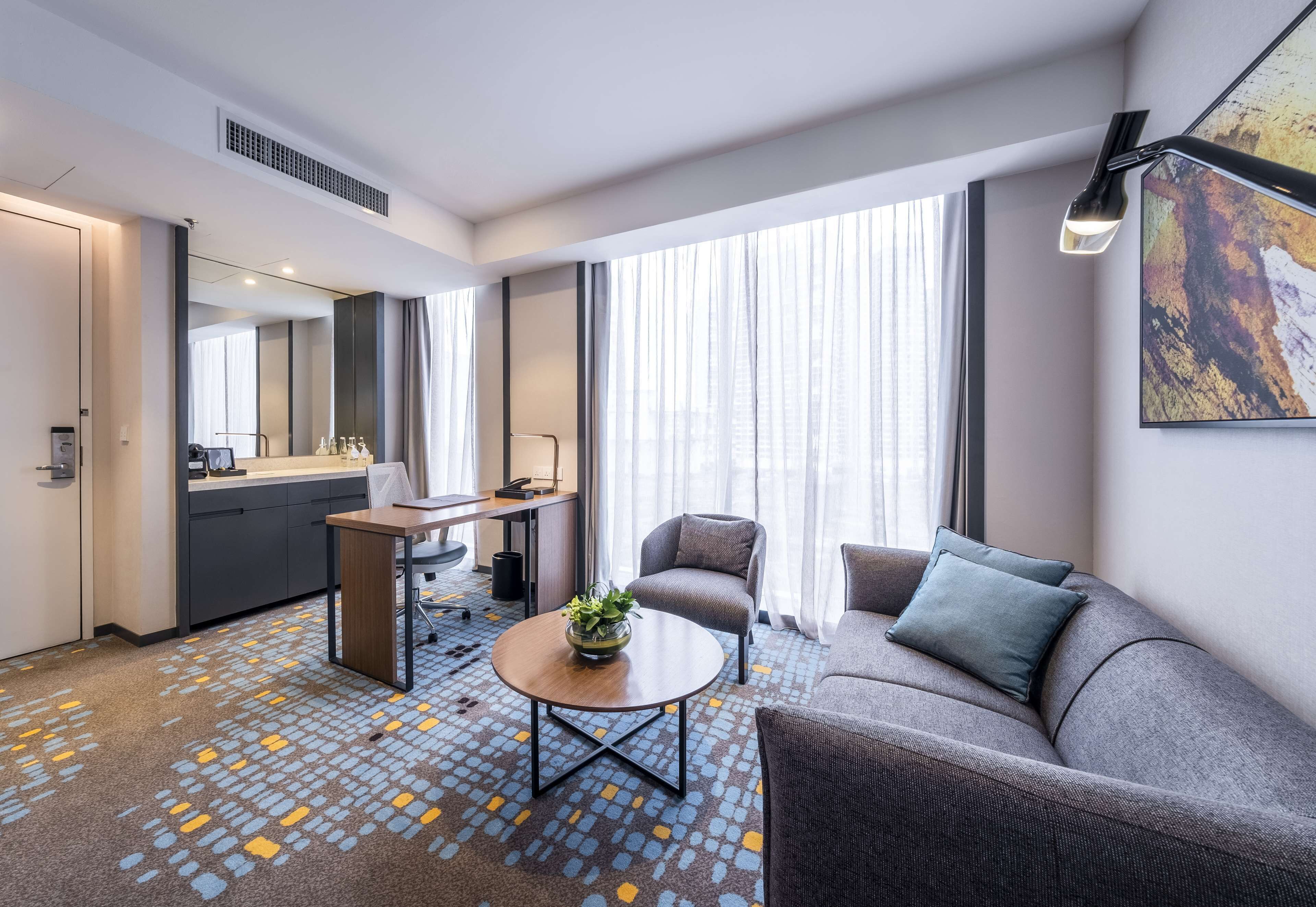 A modern hotel room with a sofa, a coffee table with a plant, a dining area with a kitchen counter, and large windows with sheer curtains.