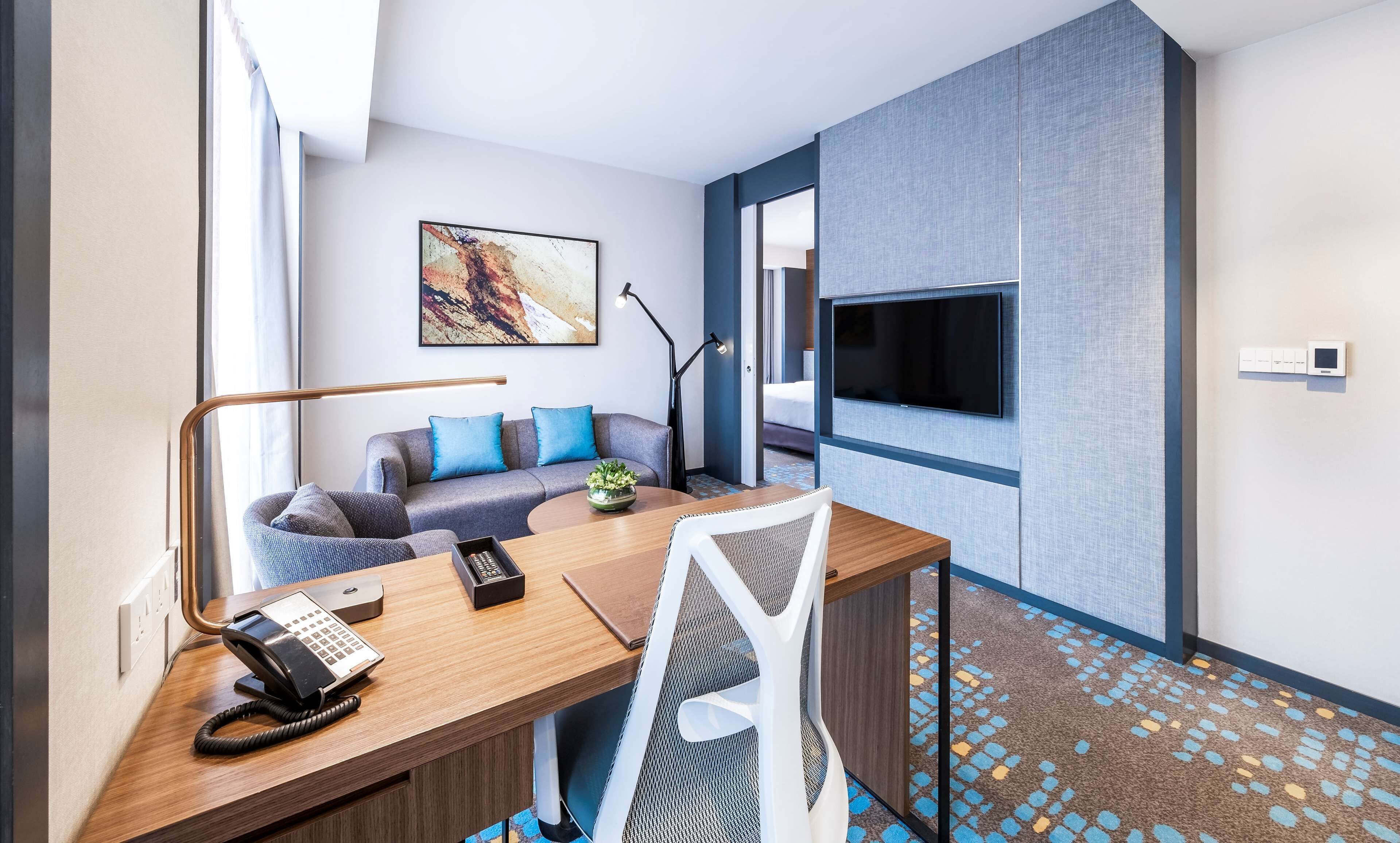 A modern hotel room with a wooden desk, a telephone, a sofa with blue cushions, a flat-screen TV mounted on the wall, and a patterned carpet.