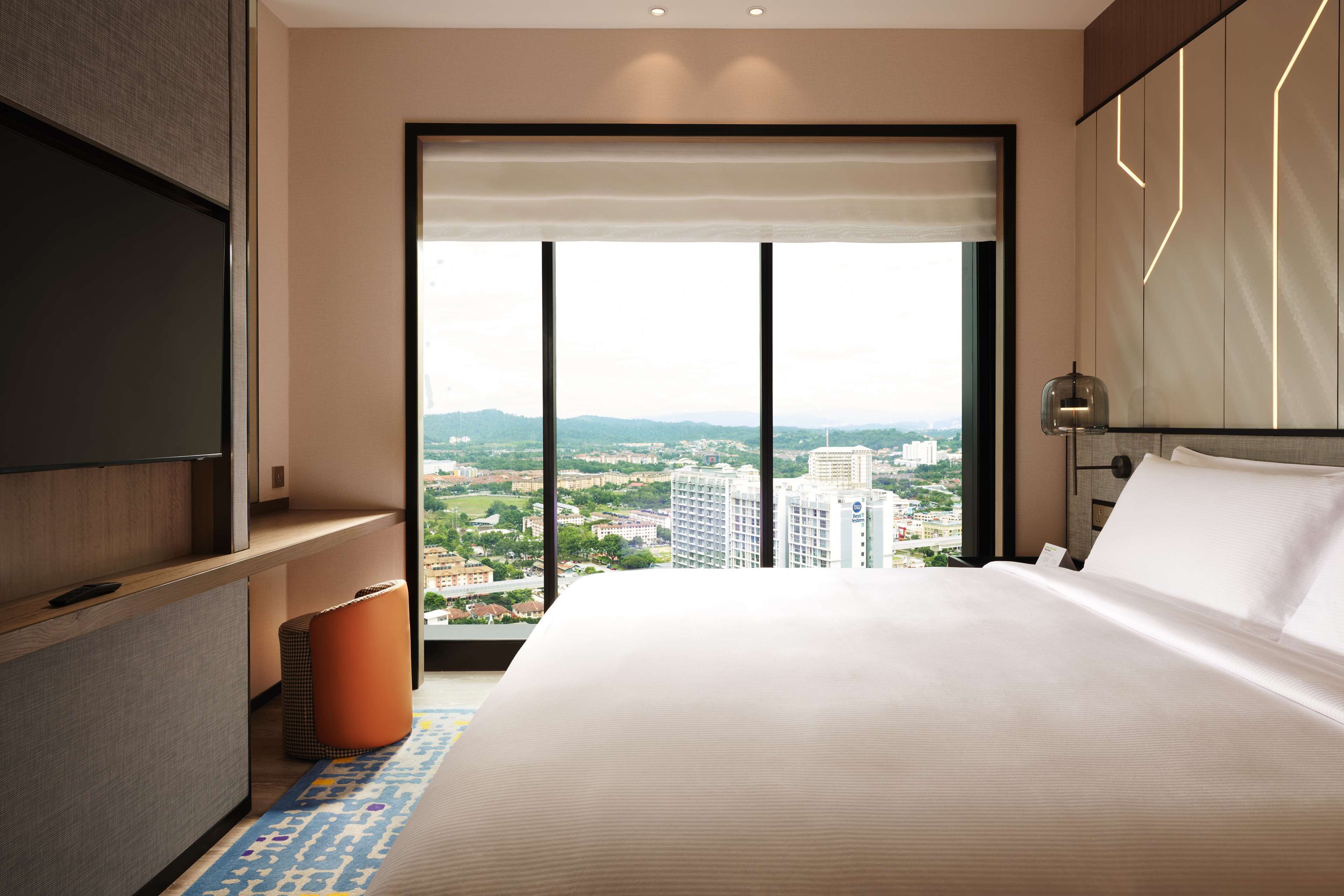 Habitación de hotel moderna con una cama grande, televisor y vista del paisaje urbano a través de ventanas de piso a techo.
