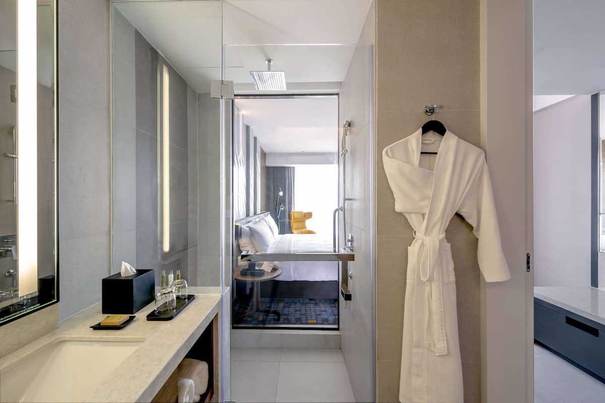 Un baño de hotel moderno con cabina de ducha de vidrio, una bata blanca colgada en la pared y una mesita de noche con artículos de tocador.