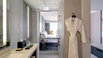A modern hotel bathroom with a glass shower enclosure, a white robe hanging on the wall, and a small bedside table with toiletries.