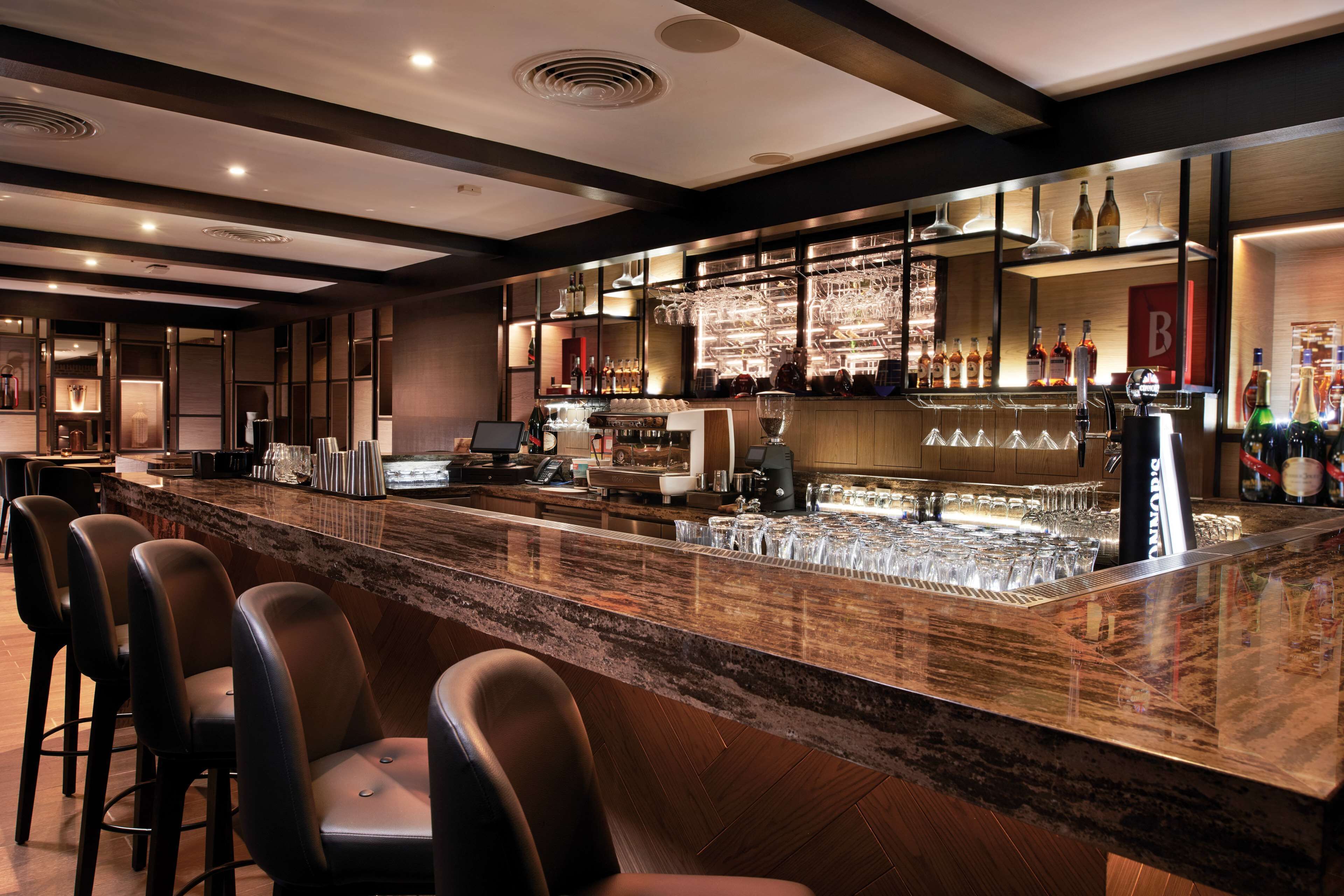 A bar area with a marble countertop, bar stools, and a well-stocked shelf with bottles and glasses.