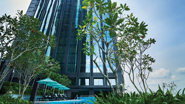 A modern hotel with a pool area, surrounded by greenery and a tall building in the background.