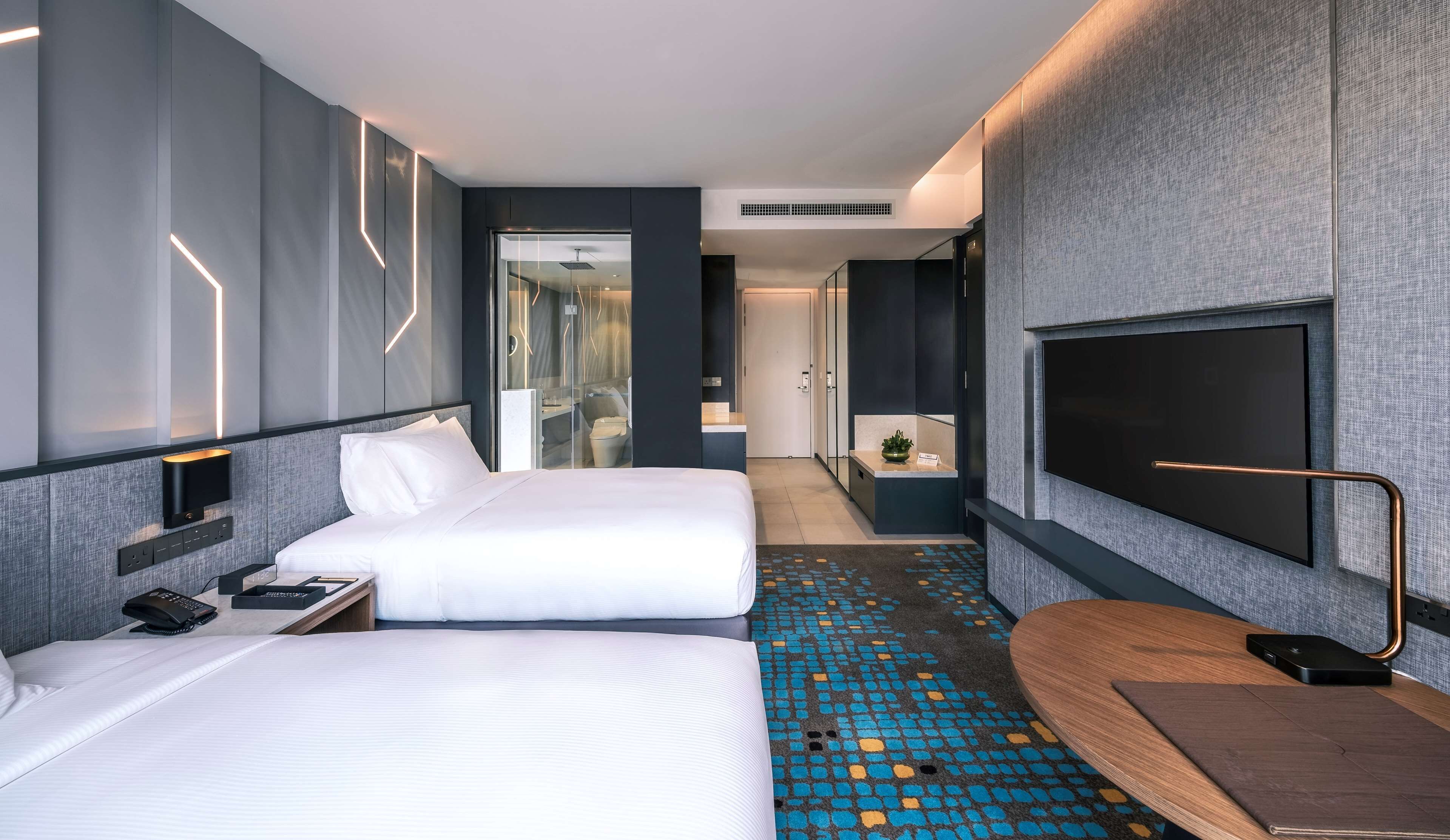 Habitación de hotel moderna con dos camas, un televisor, un escritorio y un baño visible a través de una puerta de vidrio.