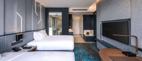 Habitación de hotel moderna con dos camas, un televisor, un escritorio y un baño visible a través de una puerta de vidrio.