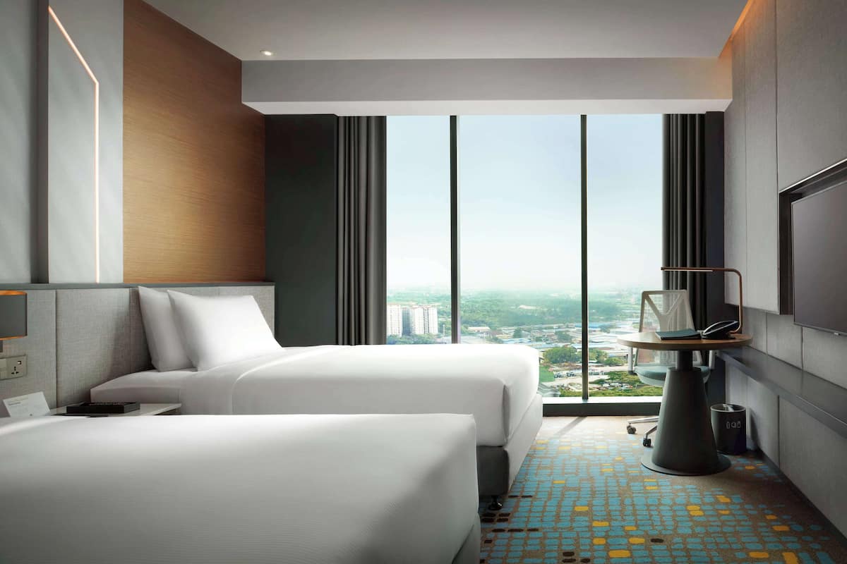 Habitación de hotel con dos camas, un ventanal con vista a la ciudad, una mesita con una silla y un cubo de basura.