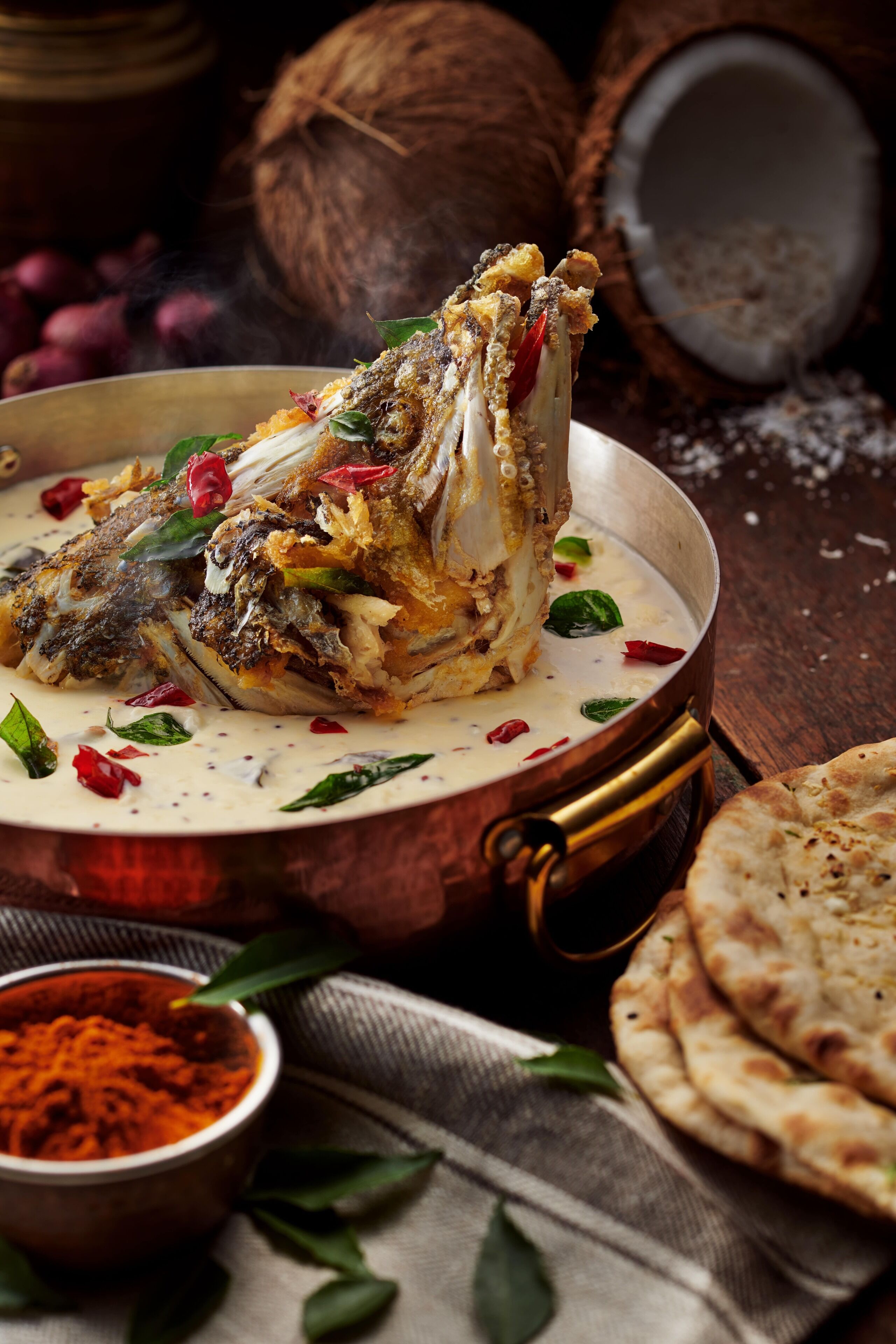 A dish of fried fish with a creamy sauce, garnished with herbs and chili, served on a copper plate.