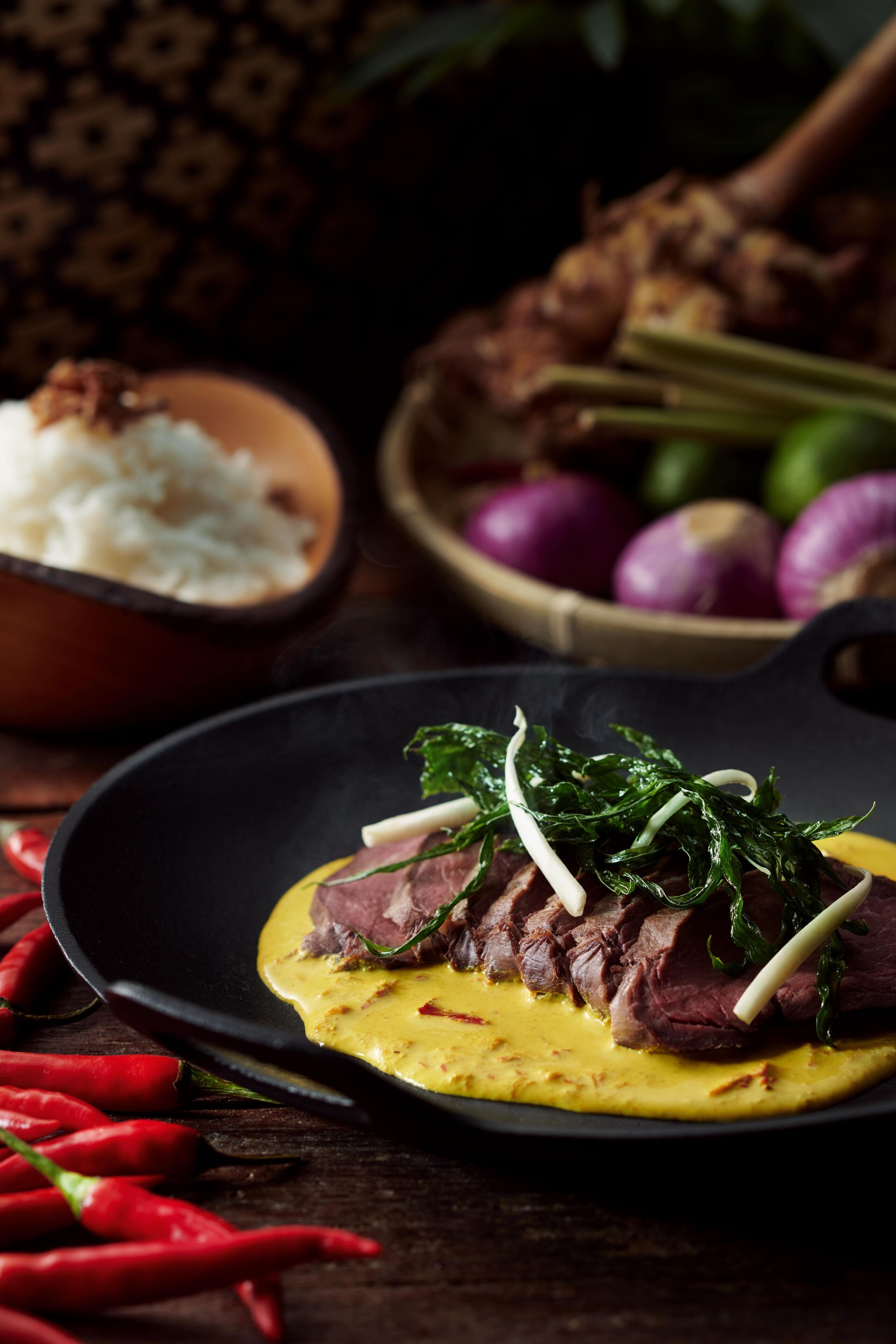 A plate of steak with greens and sauce, surrounded by red chili peppers and a bowl of rice.