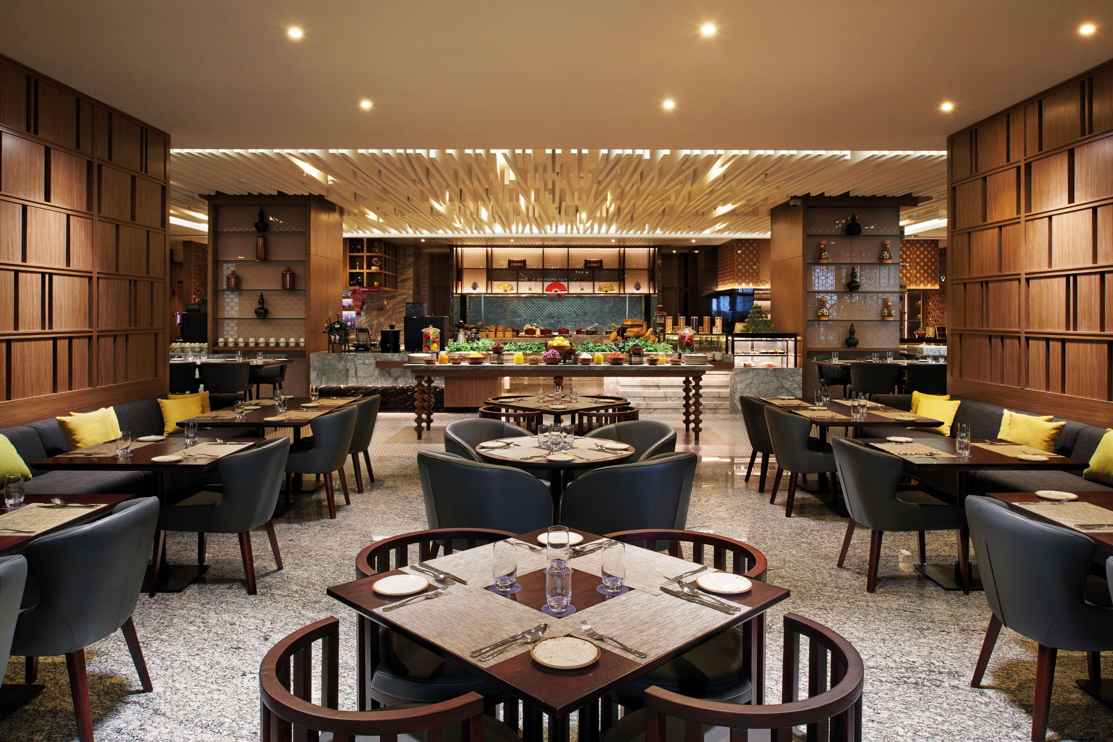 A restaurant with multiple dining areas, including round and square tables with chairs, a bar area, and a view of the kitchen in the background.