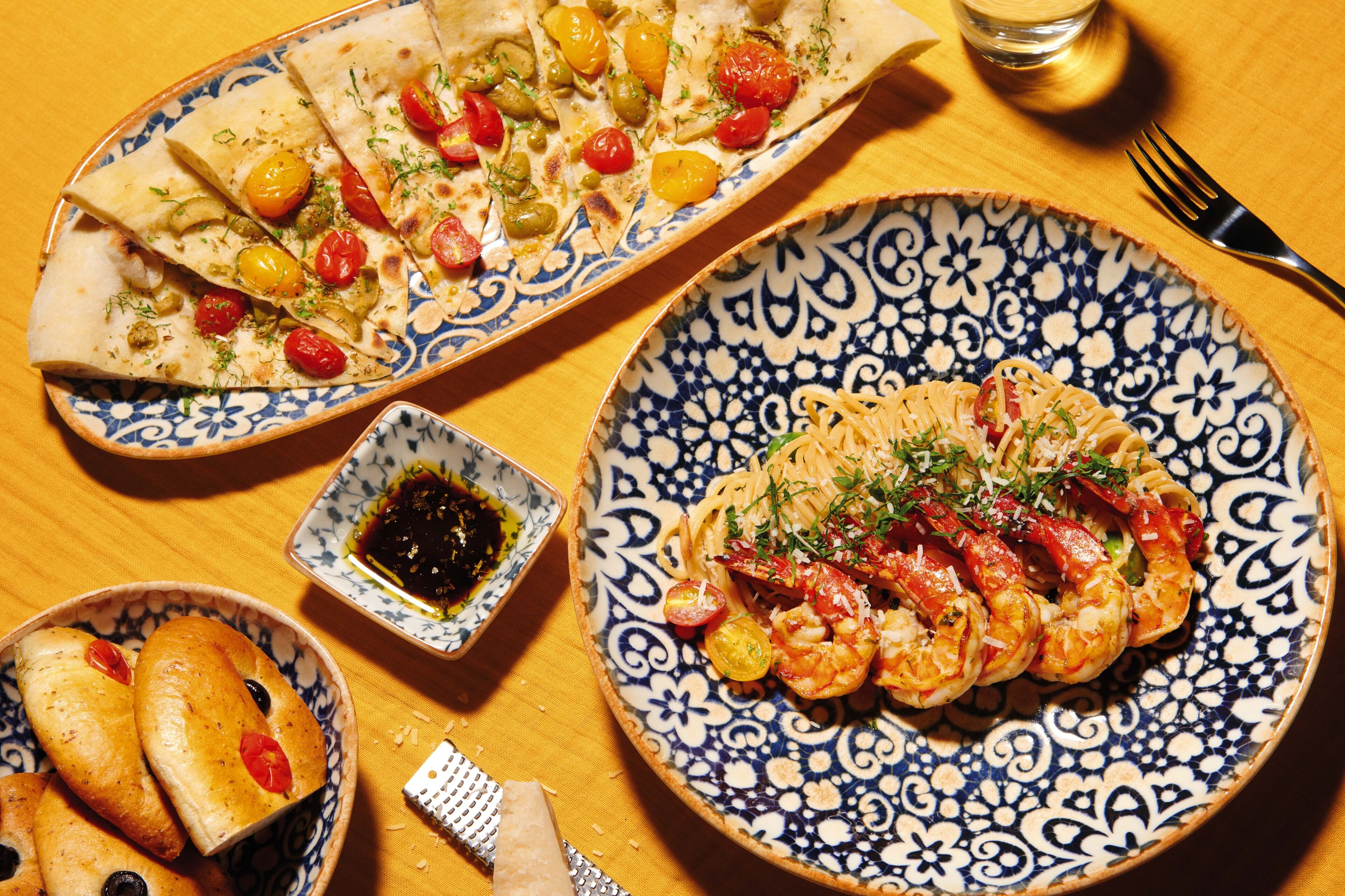 A table with a variety of dishes including pasta with shrimp, flatbread with vegetables, and bread with olives and tomatoes.
