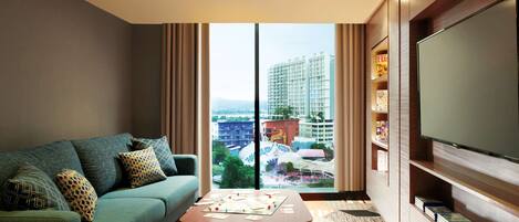 A modern hotel room with a sofa, a coffee table, a bookshelf, and a large window offering a city view.