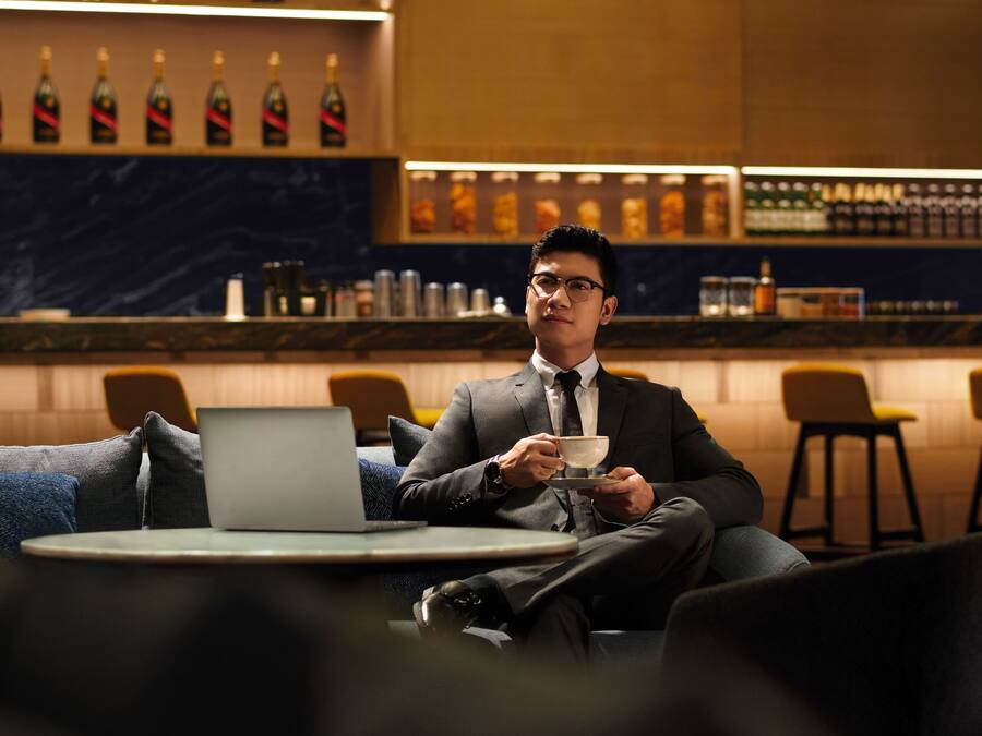 A man in a suit sitting at a table with a laptop, holding a cup.