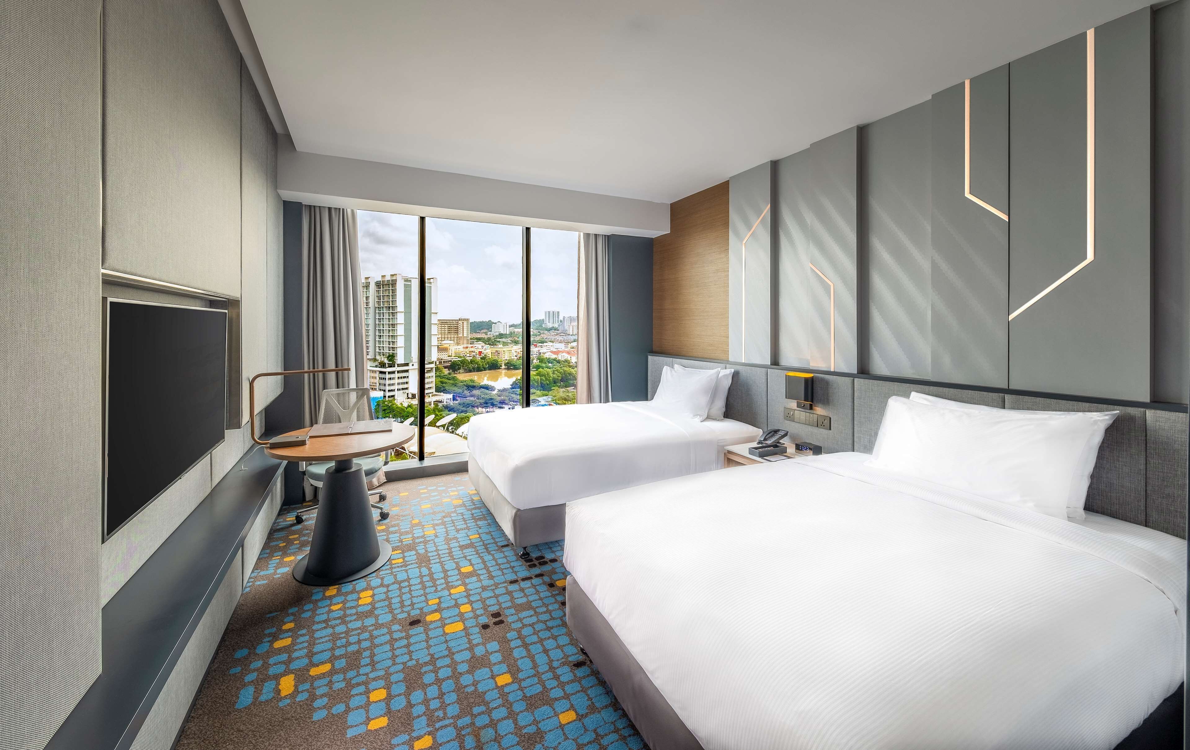 A hotel room with two beds, a desk, a chair, a TV, and a large window offering a city view.