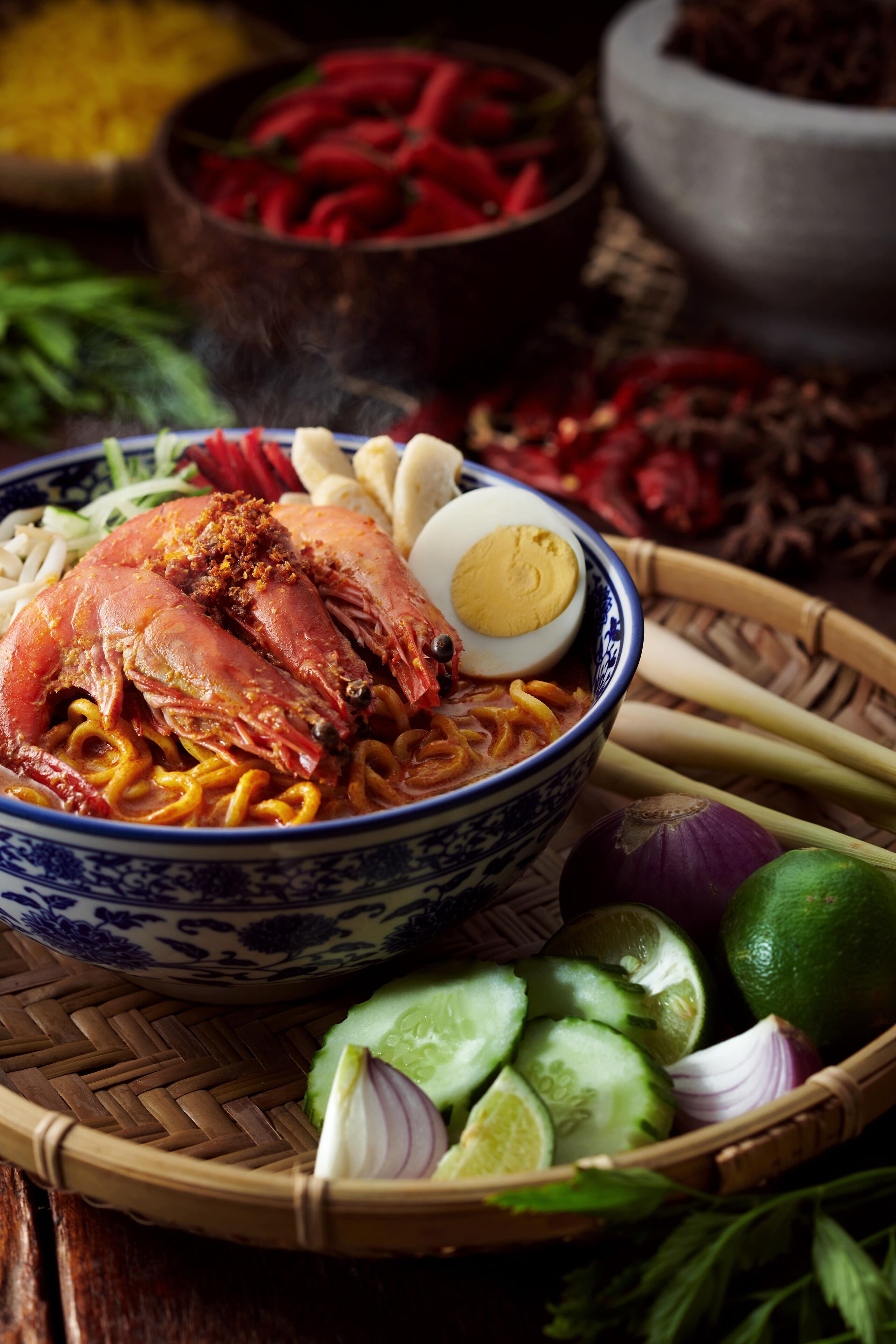Un tazón de fideos con un cangrejo y un huevo cocido, rodeado de pepinos, cebollas y limas en rodajas.