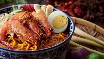 Un tazón de fideos con un cangrejo y un huevo cocido, rodeado de pepinos, cebollas y limas en rodajas.