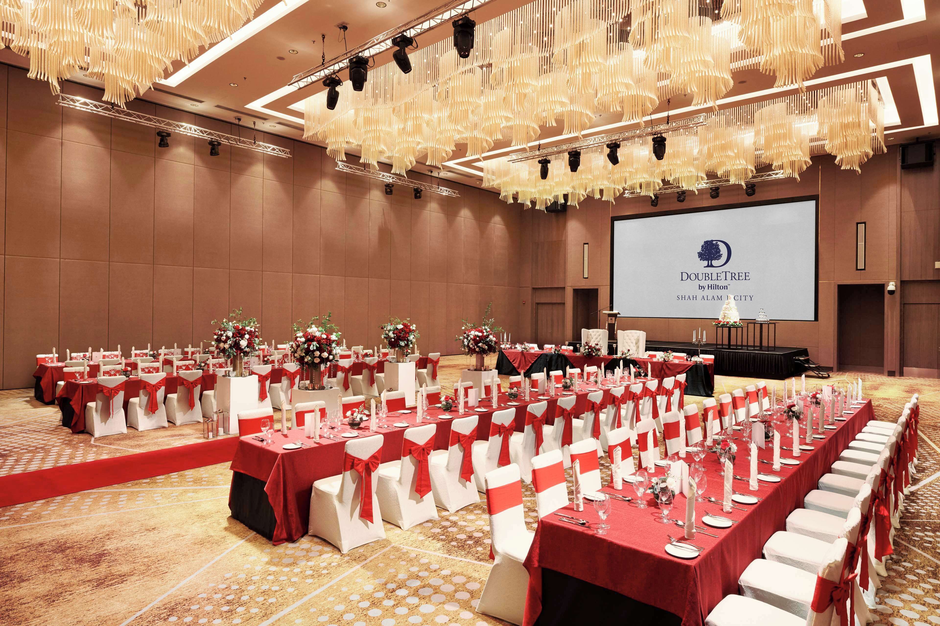 Une salle de banquet avec des rangées de tables dressées pour un événement officiel, un grand écran affichant le logo de DoubleTree by Hilton, et une scène avec un pupitre.