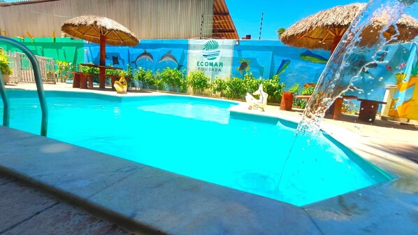 Piscina com um elemento aquático, rodeada por plantas tropicais e um mural azul com vida marinha.