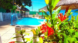 Uma piscina com cerca branca, estrutura com telhado de palha e vegetação exuberante.