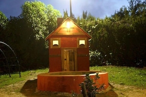 Une petite construction aux toits rouges, dotée d’une porte en bois et de deux fenêtres, et entourée de verdure.