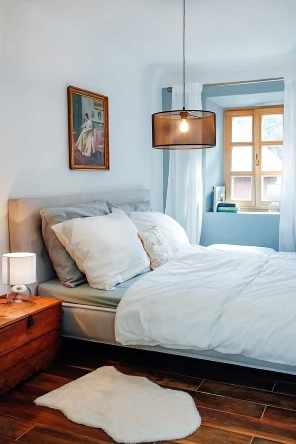 Un dormitorio con una cama, una mesita de noche de madera, una alfombra blanca, un lámpara colgante y una ventana con cortinas.