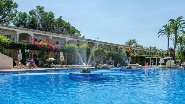 A swimming pool with a fountain, lounge chairs, and a building with arches and greenery. - Mariners Club Apartments (Alcúdia)