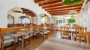A spacious dining area with wooden tables and chairs, large hanging pendant lights, and arched white walls. - Mariners Club Apartments (Alcúdia)