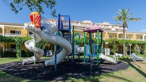 A playground with a slide and climbing structures, surrounded by a building with balconies and palm trees. - Mariners Club Apartments (Alcúdia)