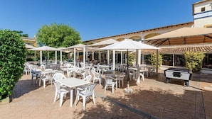 An outdoor patio area with white tables and chairs, umbrellas, and a pool table. - Mariners Club Apartments (Alcúdia)