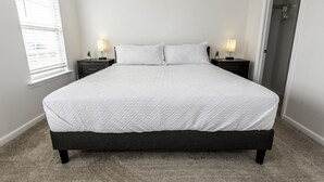 Ein ordentlich bezogenes Bett mit weißer gesteppter Tagesdecke und zwei Kissen, an beiden Seiten jeweils ein Nachttisch mit Lampe, in einem Zimmer mit Teppichboden und einer Tür rechts.
