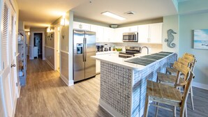 A modern kitchen with white cabinetry, a stainless steel refrigerator, a built-in microwave, and a central island with bar stools. - EMERALD OASIS IN THE HEART OF PCB!  (Panama City Beach)