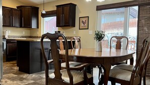 Un comedor con una mesa redonda de madera y sillas, una cocina al fondo y vistas de un edificio a través de una ventana.
